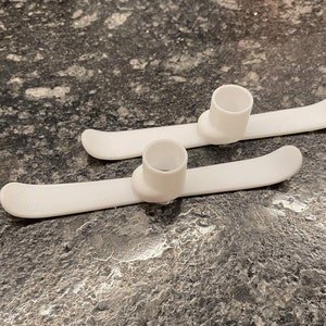 May include: Two white miniature skis with cylindrical attachments, likely for a toy or decorative item. The skis are curved at the front and have a smooth, clean appearance. The attachments are centered on the skis.
