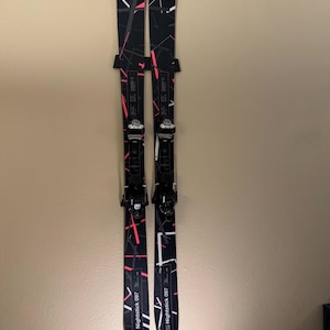 K&ouml;nnte beinhalten: Paar schwarze und wei&szlig;e Fischer Nightstick 097 Skier mit rosa und wei&szlig;en Akzenten. Die Skier sind an einer Wand montiert und zeigen ihr Design und ihre Bindungen. Die Skier haben ein geometrisches Muster.