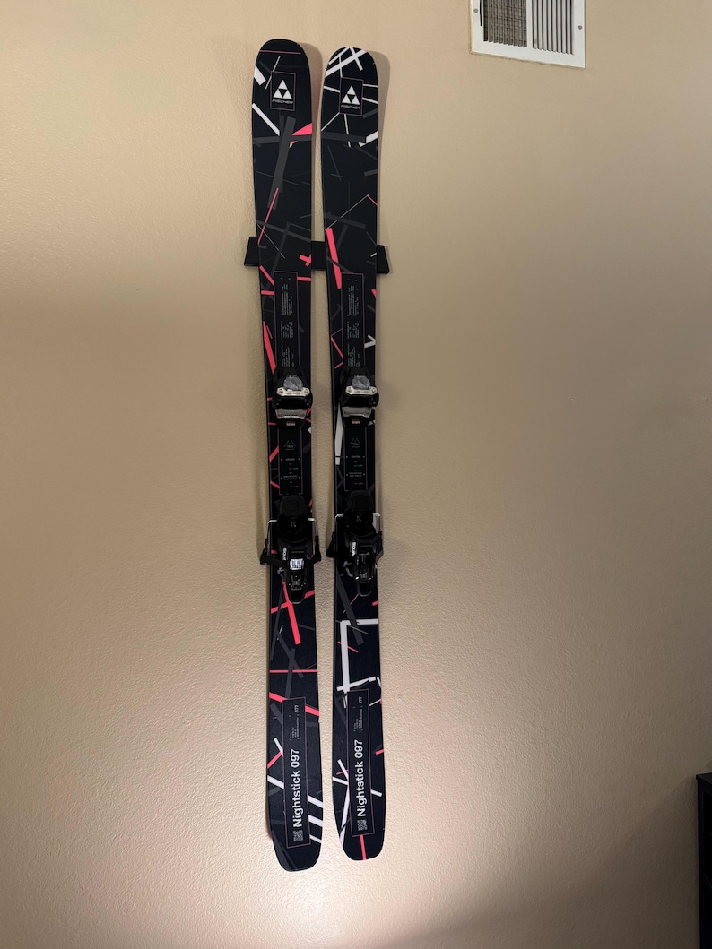 K&ouml;nnte beinhalten: Paar schwarze und wei&szlig;e Fischer Nightstick 097 Skier mit rosa und wei&szlig;en Akzenten. Die Skier sind an einer Wand montiert und zeigen ihr Design und ihre Bindungen. Die Skier haben ein geometrisches Muster.