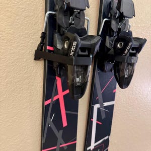 K&ouml;nnte beinhalten: Paar schwarze und rosa Ski mit schwarzen Bindungen. Die Ski haben ein geometrisches Design in Rosa und Wei&szlig;. Die Bindungen sind mit "Squire" beschriftet. Die Ski sind montiert und einsatzbereit.