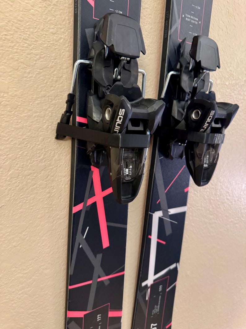 K&ouml;nnte beinhalten: Paar schwarze und rosa Ski mit schwarzen Bindungen. Die Ski haben ein geometrisches Design in Rosa und Wei&szlig;. Die Bindungen sind mit "Squire" beschriftet. Die Ski sind montiert und einsatzbereit.