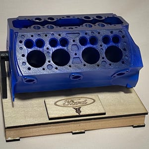 May include: A blue engine block model with multiple circular openings and a rectangular wooden base. The base features a wooden plaque with the Ford logo and V8 emblem. The model is a detailed representation of an engine component.