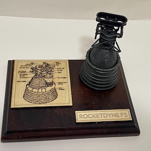 May include: A wooden plaque with a black and silver model of a Rocketdyne F1 rocket engine. The plaque has a laser-engraved diagram of the engine and the text "ROCKETDYNE F1".