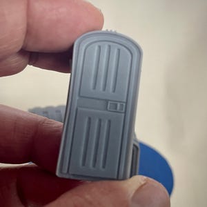 May include: A gray plastic miniature model of a portable toilet. The model has a door with a handle and vertical lines.