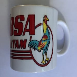 May include: White ceramic mug with the text "BSA BANTAM" in red, and a colourful rooster illustration within a red circle. The mug has a white handle, suitable for hot drinks.
