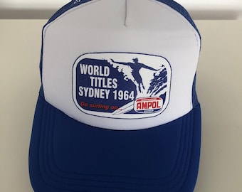 May include: A blue and white trucker hat with a patch that says "World Titles Sydney 1964" and a surfer riding a wave. The patch also says "Go surfing on..." and has the Ampol logo.