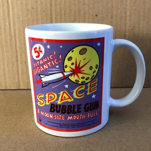 May include: White ceramic mug with a retro design featuring a rocket ship, a yellow moon, and the words "SPACE BUBBLE GUM." The mug has a purple background with the text "A moon-size mouth full!" and "Titanic! Gigantic!"