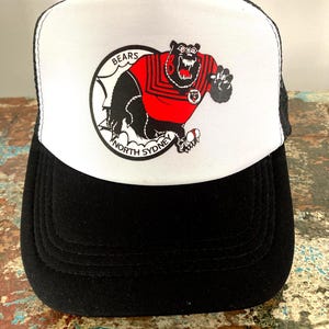 May include: Black and white trucker hat with a white front panel displaying a bear graphic in a red jersey. The bear is within a circular emblem with the words "BEARS" and "NORTH SYDNEY". The hat has a black brim and mesh back.