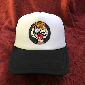 Może przedstawiać: Czarno-biała czapka typu trucker z białym panelem przednim. Posiada okrągłą naszywkę z ilustracją tygrysa i napisem "BALMAIN TIGERS". Czapka ma czarny daszek i siatkową tylną część.