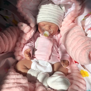 Op de afbeelding: Een realistische babypop met een roze gestreepte onesie, een witte luier en een gestreepte muts. De pop heeft een fopspeen en is genesteld in een roze, zachte capuchondeken. De deken heeft een zacht, getextureerd uiterlijk.