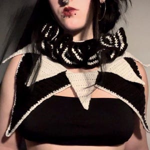 May include: A black and white crocheted collar with a triangular shape. The collar is worn around the neck and has a pointed edge.
