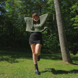 May include: A woman wearing a green crocheted sweater with a black mini skirt and black boots. She is standing in a forest with green trees and a large tree trunk behind her.