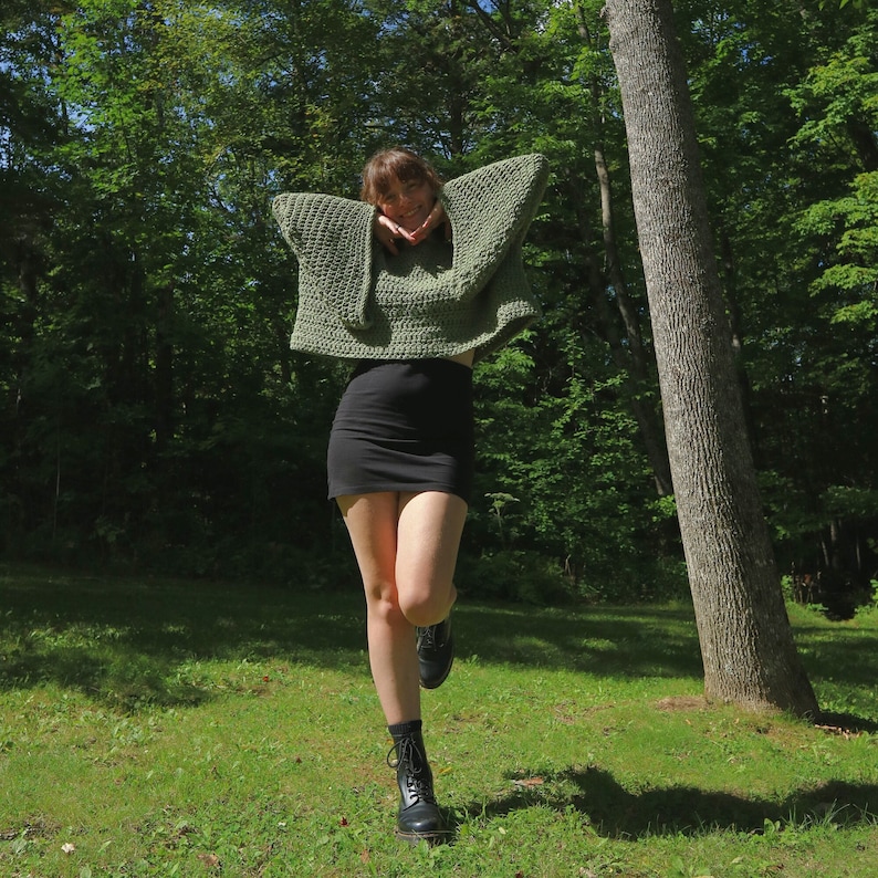 May include: A woman wearing a green crocheted sweater with a black mini skirt and black boots. She is standing in a forest with green trees and a large tree trunk behind her.