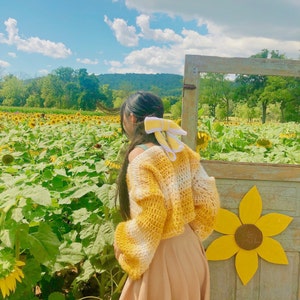 May include: A woman wearing a yellow and white crocheted sweater with a yellow bow in her hair stands in a field of sunflowers.