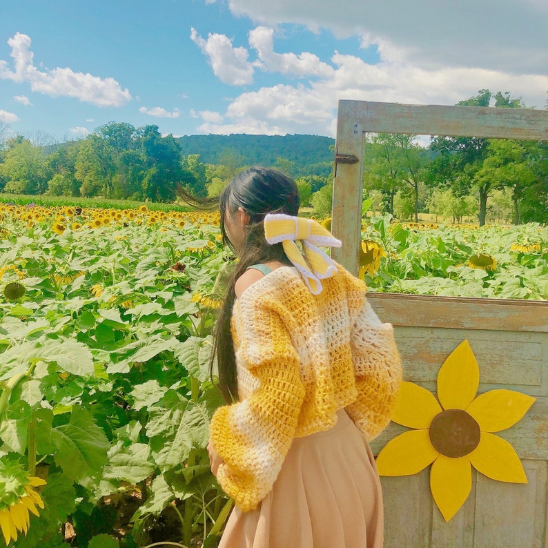 May include: A woman wearing a yellow and white crocheted sweater with a yellow bow in her hair stands in a field of sunflowers.