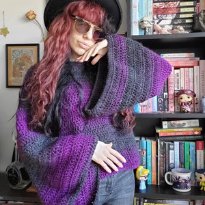 May include: A purple and black striped crocheted sweater with long sleeves and a wide, loose fit. The sweater is worn by a person with long, wavy, pink hair and a black hat.