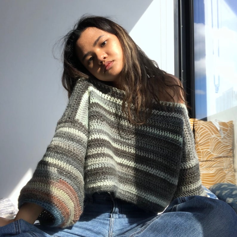 May include: A woman wearing a striped, off-the-shoulder crochet sweater with long sleeves and a rainbow stripe detail on the cuffs. She is wearing blue jeans.