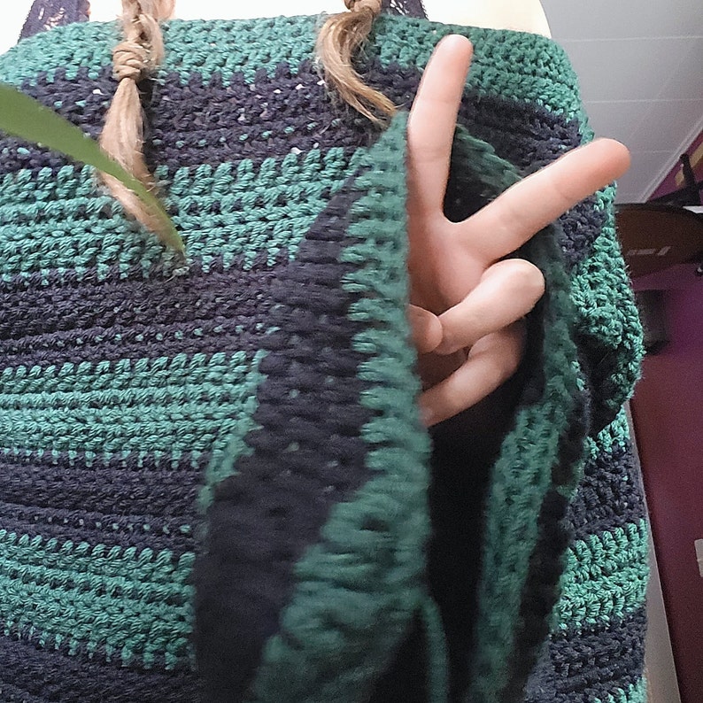 May include: A green and black crocheted shawl with a fringe border. The shawl is draped over a person's arm and hand.