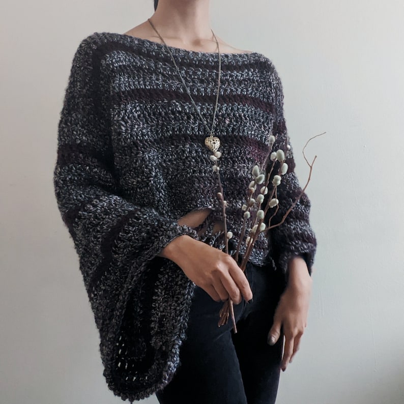 May include: A dark gray and purple striped crocheted sweater with long sleeves and a loose fit. The sweater has a wide neckline and a delicate silver heart pendant necklace.
