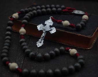 May include: A rosary with dark gray beads, cream-colored accent beads, and a silver crucifix. The beads are strung on red cord. The rosary rests on a dark, leather-bound book.