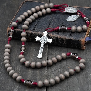 May include: A rosary with gray beads, red cord, and a silver crucifix. The rosary is draped over a dark, leather-bound book with a blue ribbon bookmark. Two silver medallions are also visible.