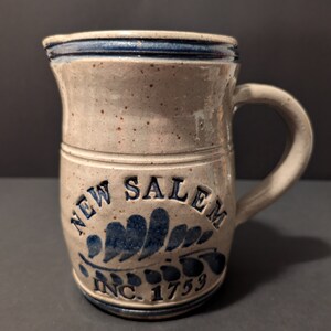 May include: A gray stoneware pitcher with a blue glaze and a blue and white design that says "NEW SALEM INC. 1753".