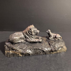 May include: A dark-toned sculpture depicting a lion and a lamb resting on a textured base. The lion is lying down, with detailed fur and mane. The lamb is positioned nearby, also in a resting pose. The base has a rough, stone-like appearance.