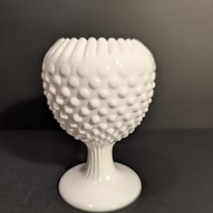May include: A white hobnail glass vase with a round base and a stem that flares out to support the bowl. The bowl has a textured surface of small, raised bumps. The rim of the vase is scalloped.