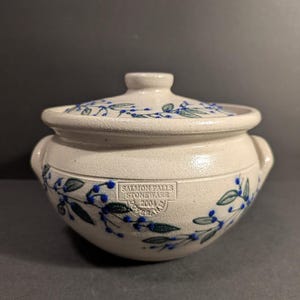 May include: A cream-colored stoneware pot with a matching lid and two side handles. The exterior features a hand-painted pattern of green leaves and small blue berries. A stamp on the front reads Salmon Falls Stoneware 2004 Dover NH.