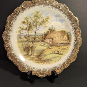 May include: Decorative plate with a rural landscape scene. The plate features a thatched-roof cottage, sheep, and a tree. The rim is gold-colored with a scalloped edge.
