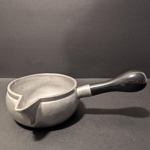 May include: A small, silver-colored saucepan with a black handle. The saucepan has a spout on the side for pouring.