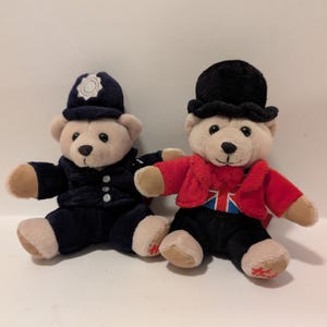May include: Two plush teddy bears dressed in British themed outfits. One bear is wearing a blue police officer uniform with a black hat and the other bear is wearing a red jacket with a Union Jack design and a black top hat.