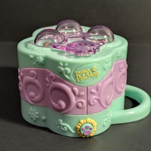 May include: A mint green plastic Littlest Pet Shop toy container with a purple band featuring embossed swirls. The top has three clear dome-shaped windows and a purple flower-shaped dial. A yellow paw print emblem is on the front, and a handle is on the side.