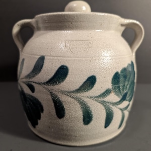 May include: A beige stoneware crock with two side handles and a matching lid. The exterior features a hand-painted dark teal floral vine pattern and a stamped maker's mark that reads Salmon Falls Stoneware 1993 Dover NH on the upper front.