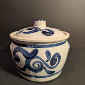 May include: A small, ceramic lidded pot with a cream-colored base and lid. The pot is adorned with blue, swirling, hand-painted designs. The lid has a small knob for easy removal. The pot has two small handles on either side.