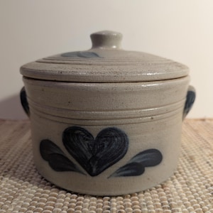 May include: A stoneware lidded pot with a heart design. The pot is light beige with a dark blue heart and leaf accents. It has two small handles and a knob on the lid.