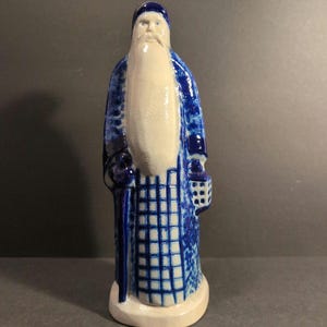 May include: A ceramic figurine of a bearded figure wearing a long blue and white patterned robe, a matching blue cap, and holding a staff in one hand and a small lantern in the other, standing on a circular base with a glossy finish.