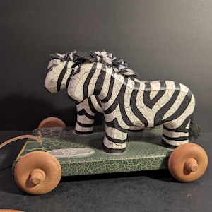 May include: A vintage wooden pull toy featuring a zebra with black and white stripes. The zebra stands on a green base with wooden wheels and a leather pull cord. The toy has a distressed, crackled finish, giving it a classic, antique appearance.
