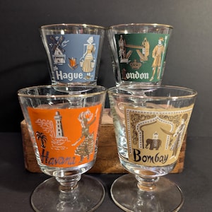 May include: Four vintage shot glasses with gold rims. Each glass features a different city: Hague, London, Havana, and Bombay. The glasses have colorful illustrations and city names printed on them.