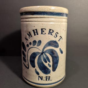 May include: A cylindrical stoneware jar with a beige base and blue floral design. The word "AMHERST" is arched across the center, with "N.H." below. The rim and base have blue bands.