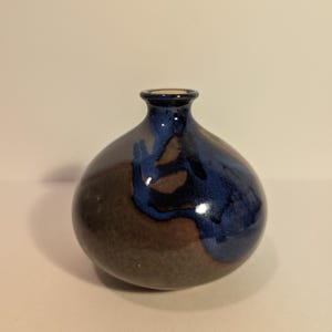 May include: A small, round ceramic vase with a narrow neck. The vase features a glossy finish with a color scheme of brown, blue, and a touch of beige at the rim. The blue paint appears to drip down the sides.