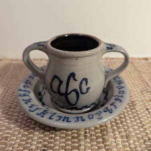 May include: A small, gray ceramic cup with two handles, sitting on a matching saucer. The cup and saucer are decorated with blue lettering and designs. The cup has the letters "abc" in blue. The saucer has the alphabet around the edge.