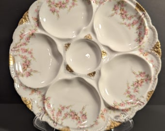 May include: A white porcelain oyster plate featuring five outer scalloped wells and one central circular well. The plate is decorated with delicate pink floral sprays and gold gilded accents along the scalloped edges and center.