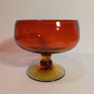 May include: A vintage-style, amber-colored glass goblet. The bowl is a deep orange, transitioning to a lighter amber at the base. The goblet has a rounded bowl, a short stem, and a circular base. The glass appears to be smooth and reflective.