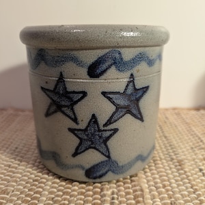 May include: A stoneware crock with a light gray base and blue star and wave designs. The cylindrical container has a rounded rim and is likely used for storage. The stars are hand-painted, adding to its rustic charm.