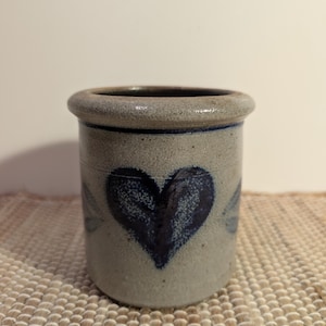 May include: A stoneware crock with a dark blue heart design. The cylindrical container has a rounded rim and a light gray background. The heart is centered on the front, with a few blue leaf-like accents. The crock is likely for kitchen use.