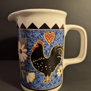 May include: White ceramic creamer with a blue speckled background featuring a black rooster, daisies, and a red heart. The top rim has a black and white geometric pattern. It has a curved spout and a square handle.