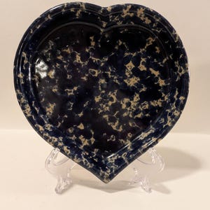 May include: A heart-shaped ceramic plate with a dark blue speckled pattern and a light beige background. The plate is displayed on a clear plastic stand, showcasing its unique shape and design.