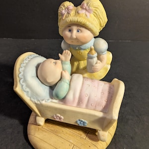 May include: A ceramic figurine featuring a baby in a crib and a doll-like figure holding a bottle. The baby is in a light blue and pink crib with floral accents. The doll figure has blonde hair and wears a yellow dress.
