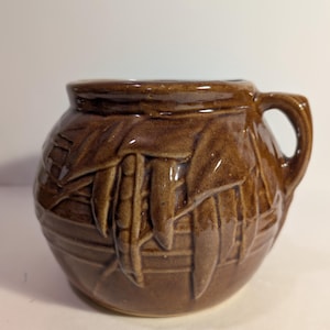 May include: A brown ceramic pot with a handle, featuring a textured design of leaves and pods. The pot has a rounded shape and a glossy finish, suitable for kitchen or decorative use.